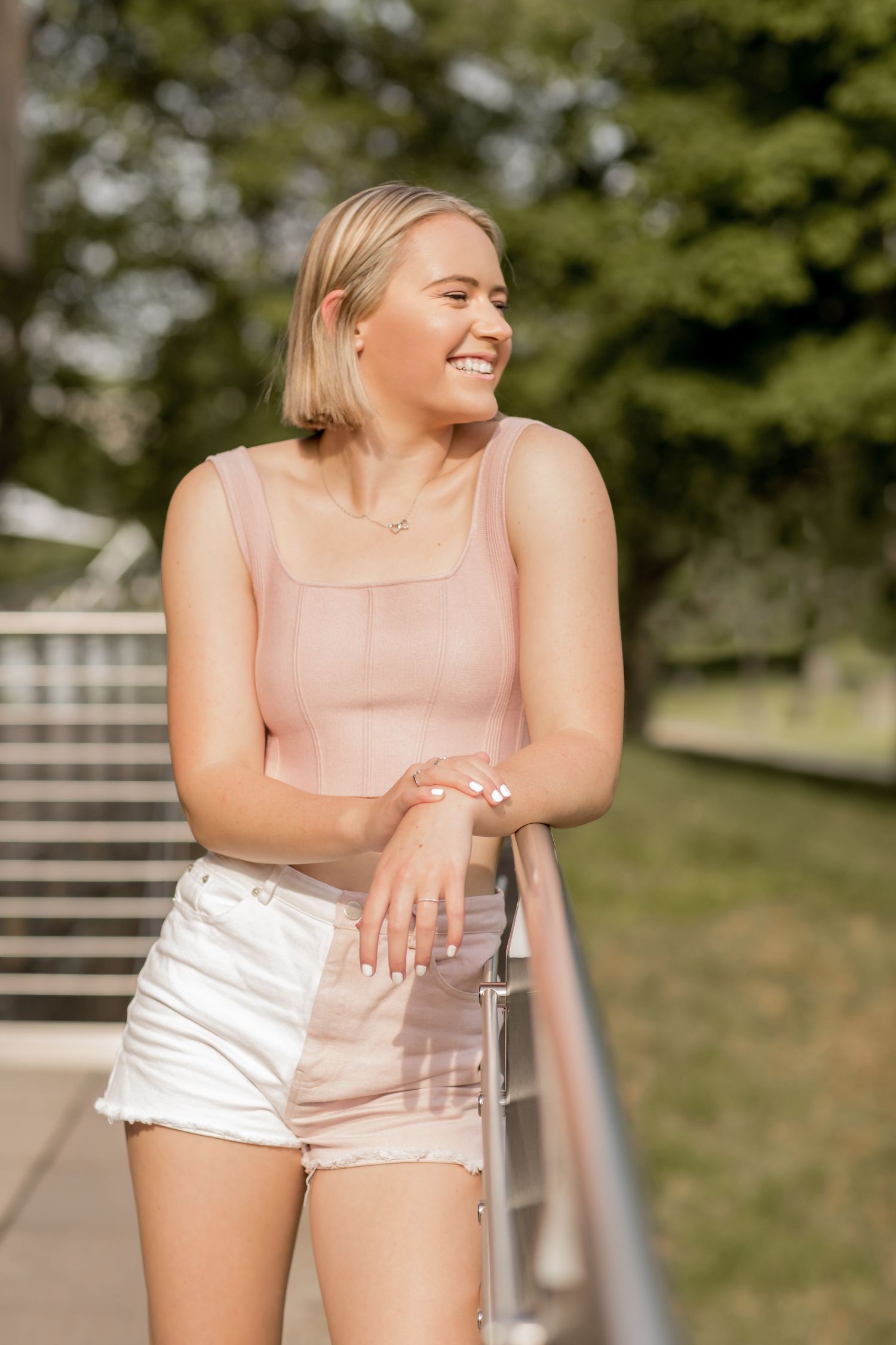 Blonde woman in pink tank top and white shorts laughing while leaning on a railing in a sunny park.