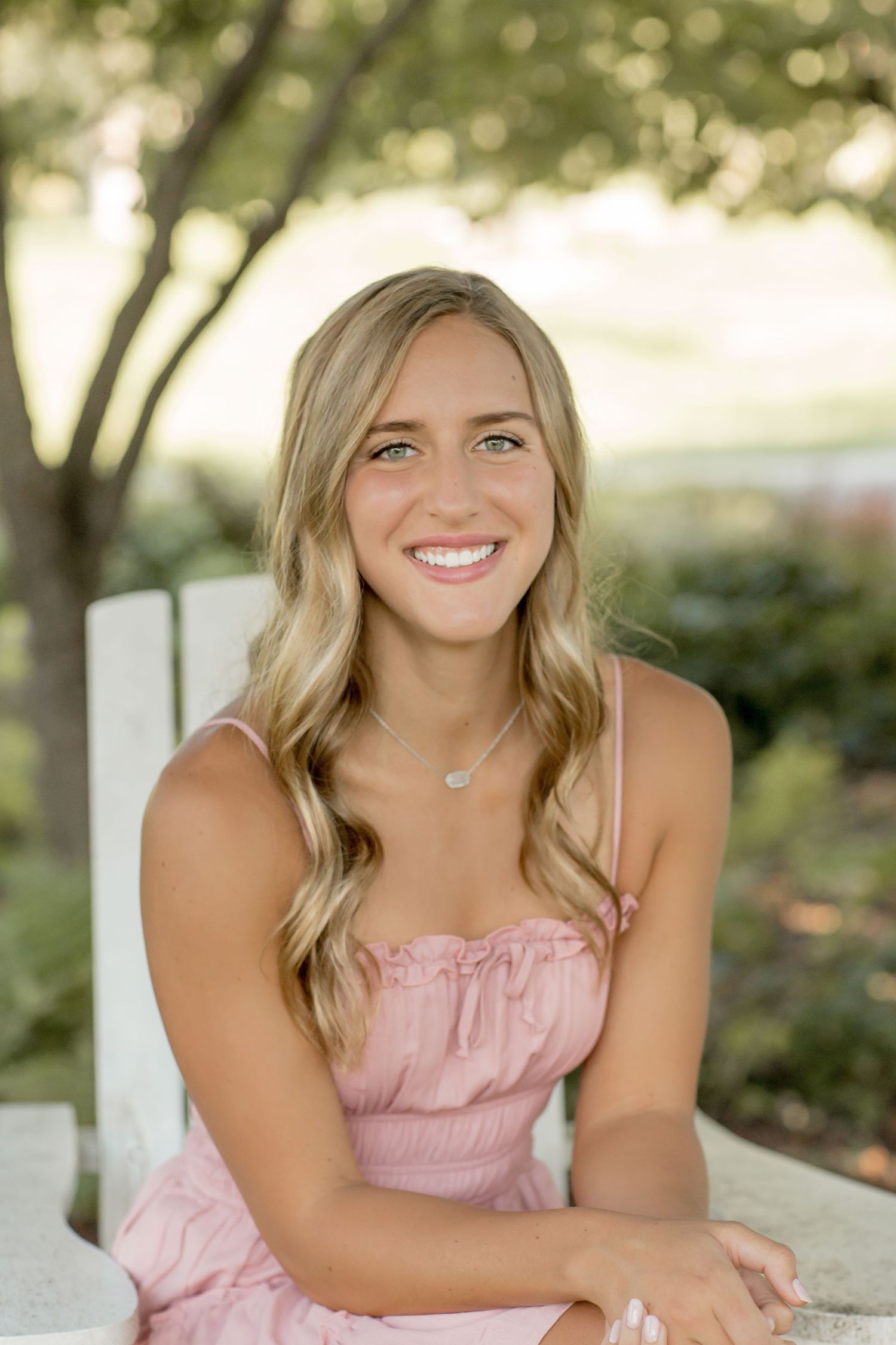 Young blonde woman in pink sundress smiling brightly while seated outdoors with lush green trees behind her.