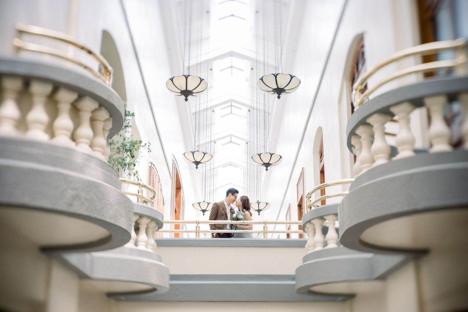 Architectural wedding photo taken in white hallway with ornate balconies and chandeliers.