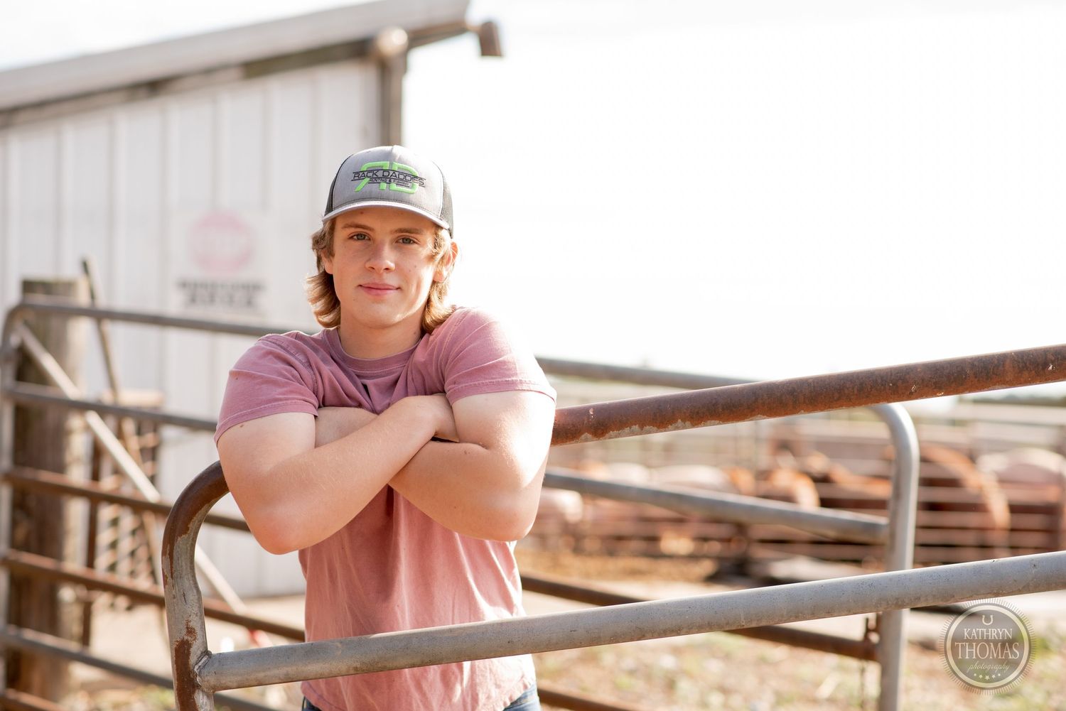 Farm worker in baseball cap and pink shirt leans on metal railing at sunset.