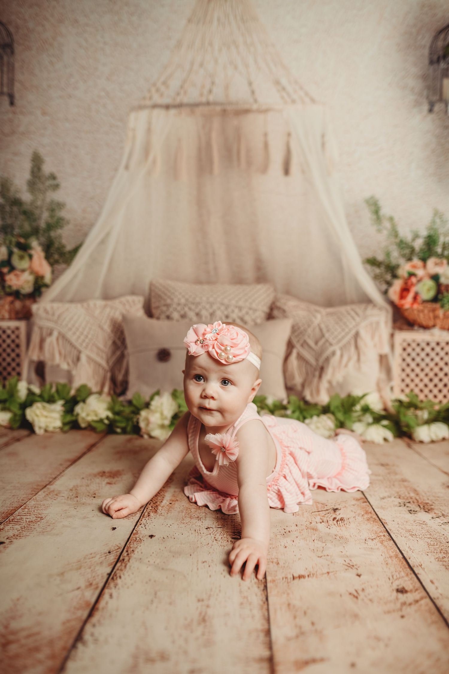 Baby crawling on wooden floor with macrame backdrop and pink floral decorations.
