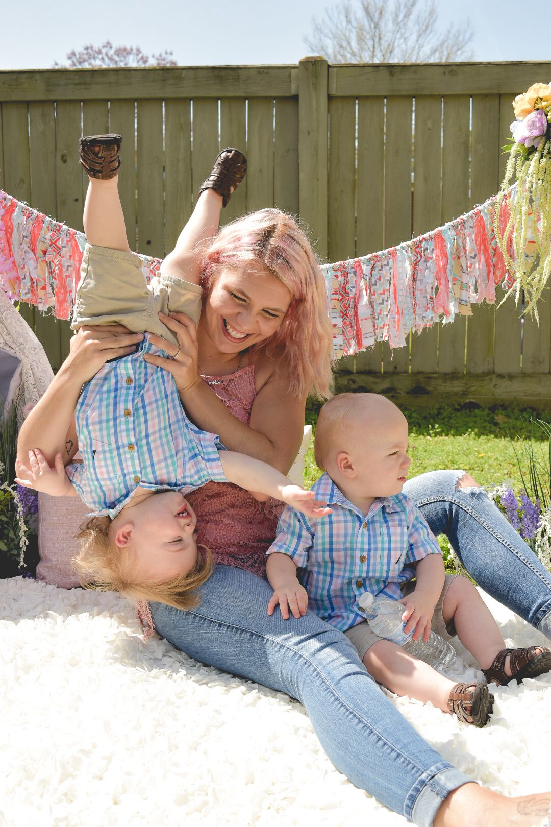 Parent playing with children on blanket under decorative banner in backyard.