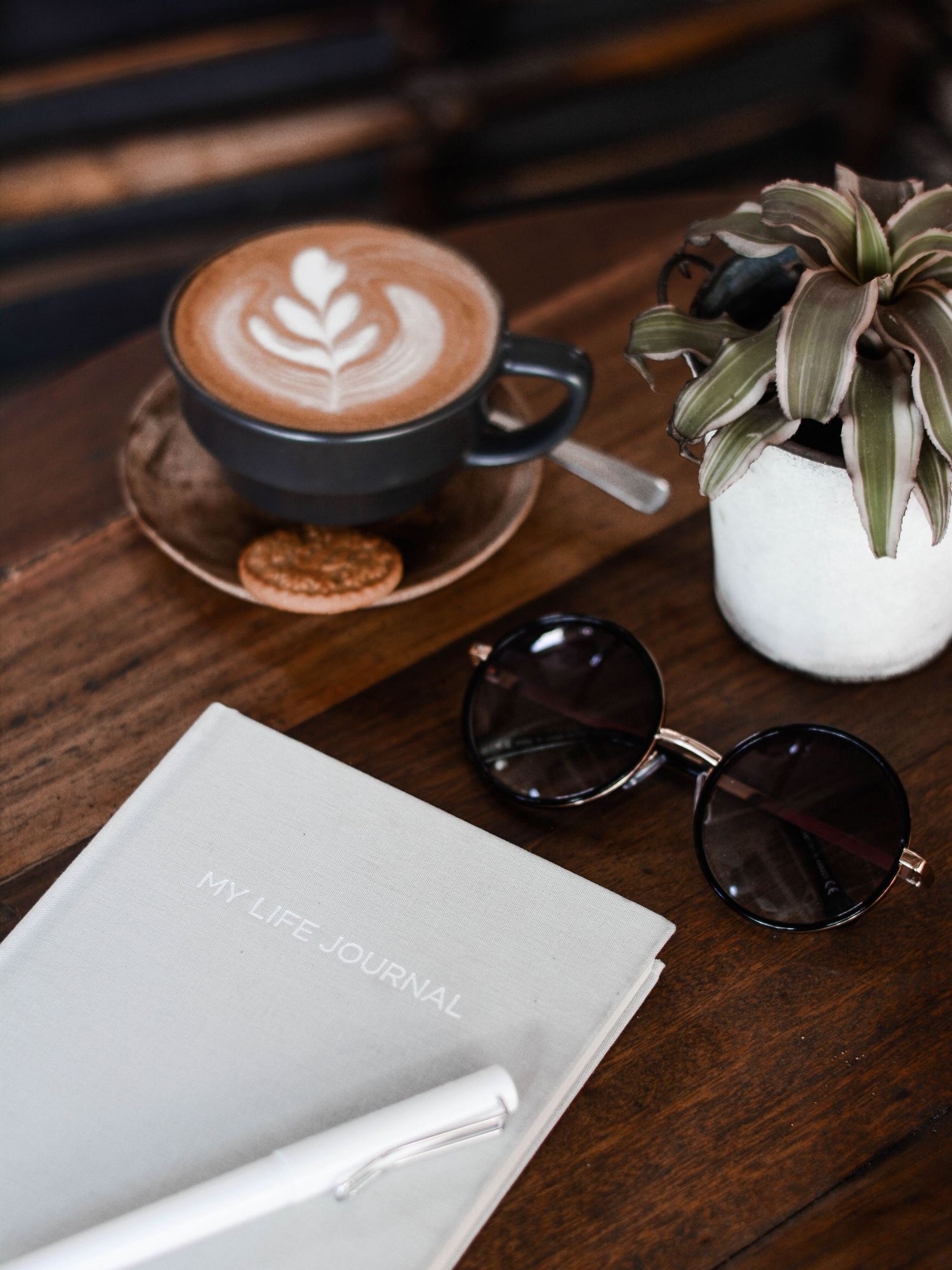 Coffee cup with latte art next to sunglasses, journal and air plant on wooden table.