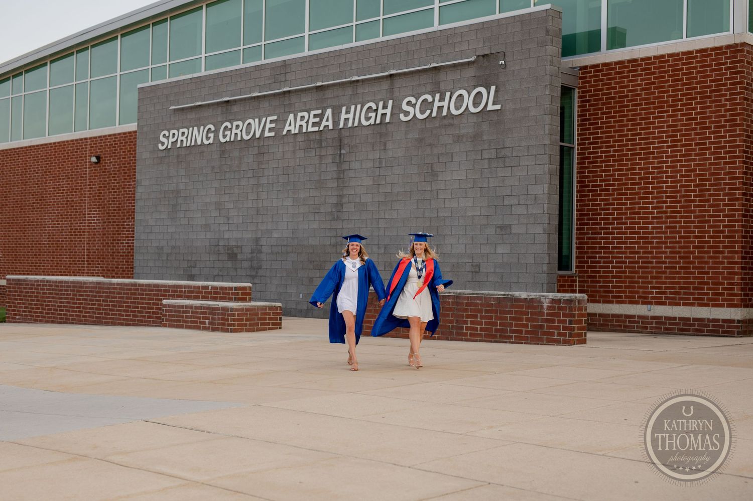 Two graduates walk together in blue caps and gowns outside their high school.