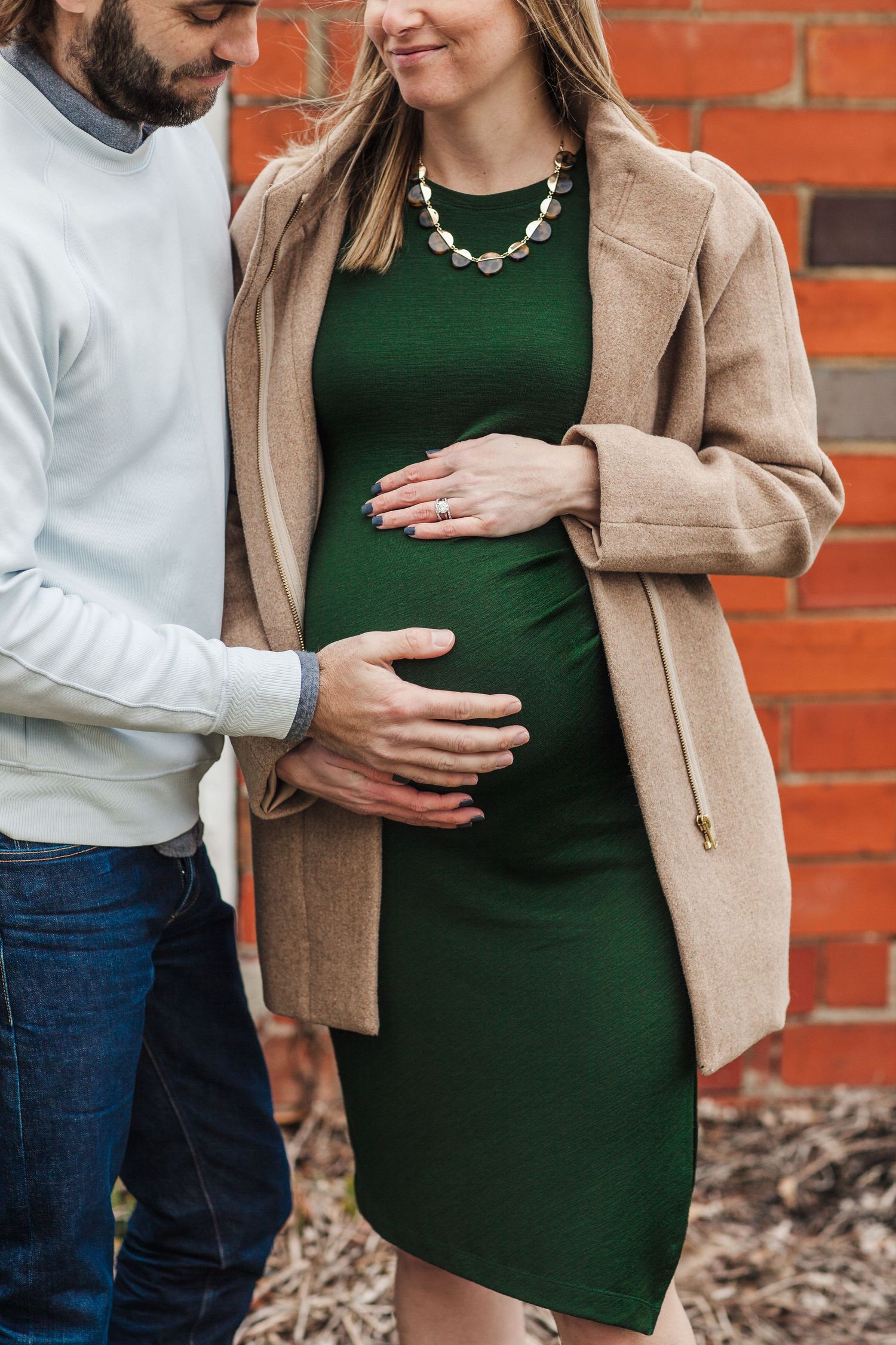 A tender maternity moment captured against a rustic brick wall with a camel coat and green dress.
