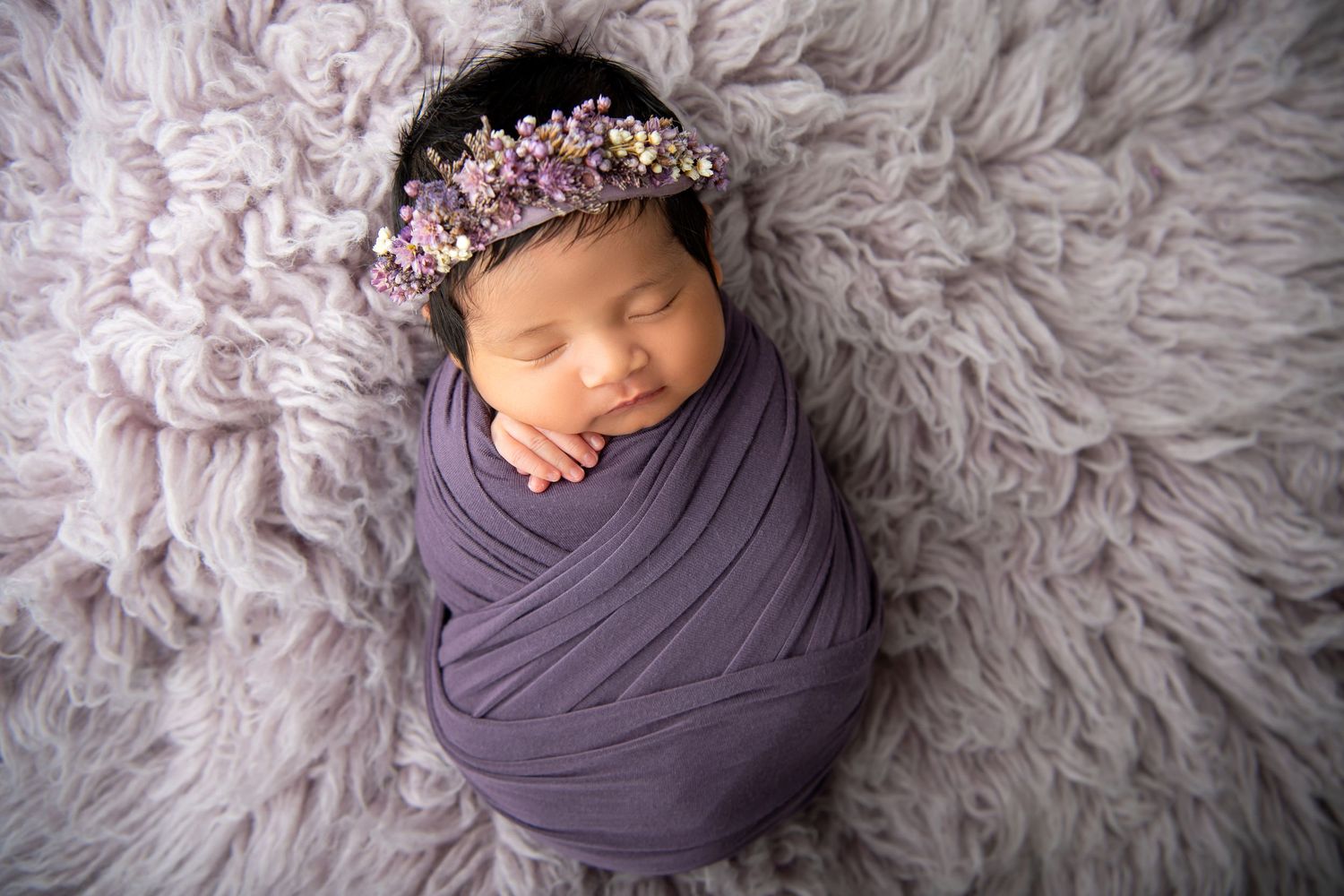 Sleeping newborn wrapped in purple swaddle with floral crown on textured gray blanket.
