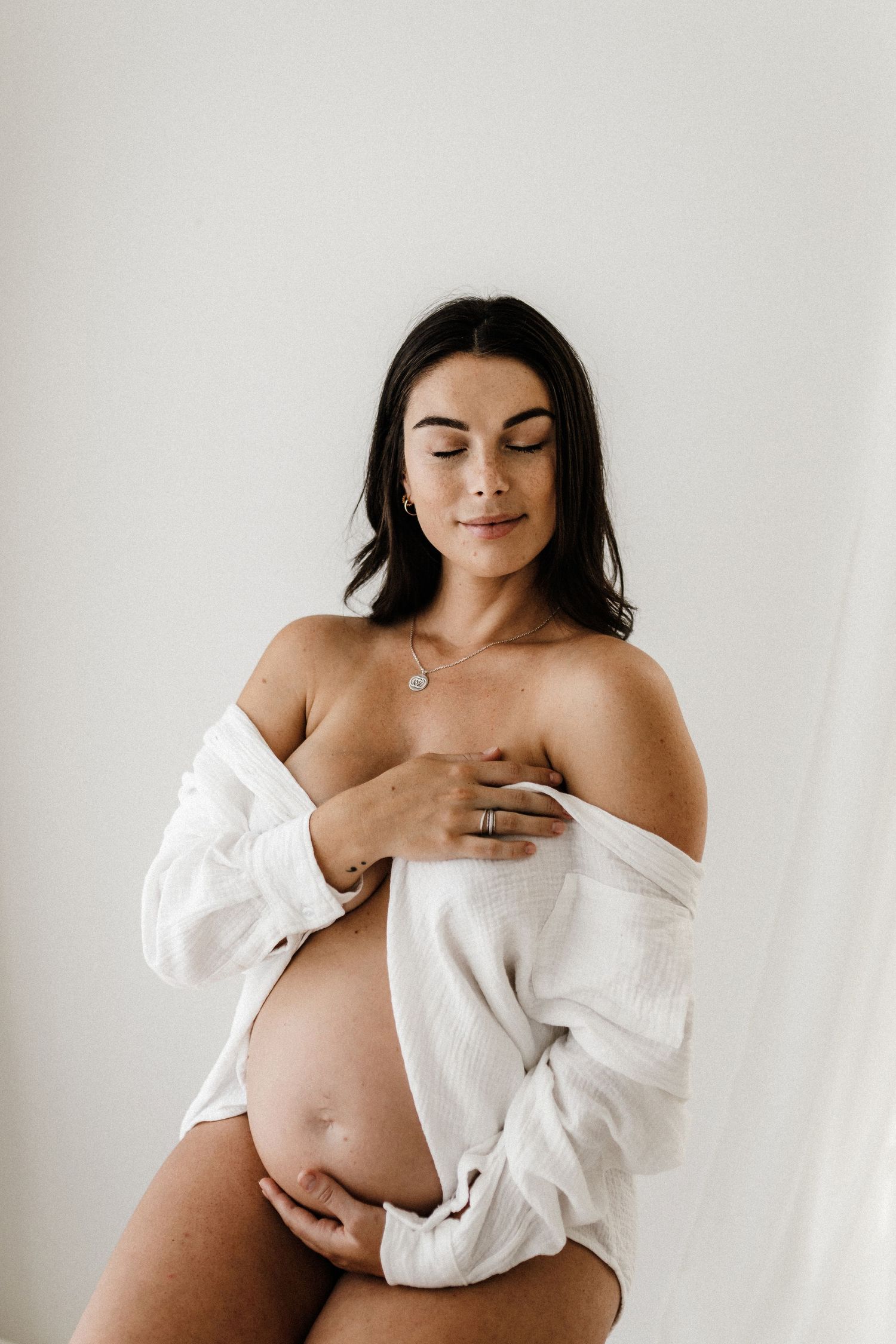 Expecting mother in white shirt poses intimately against white backdrop in maternity photoshoot.