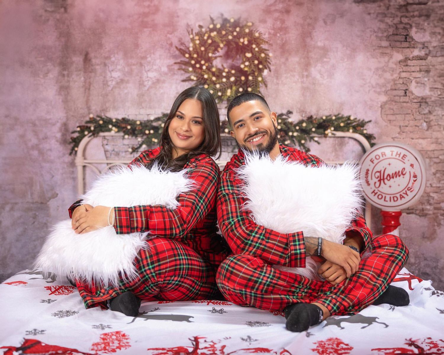 Couple in matching plaid pajamas pose on white fur with holiday decor.