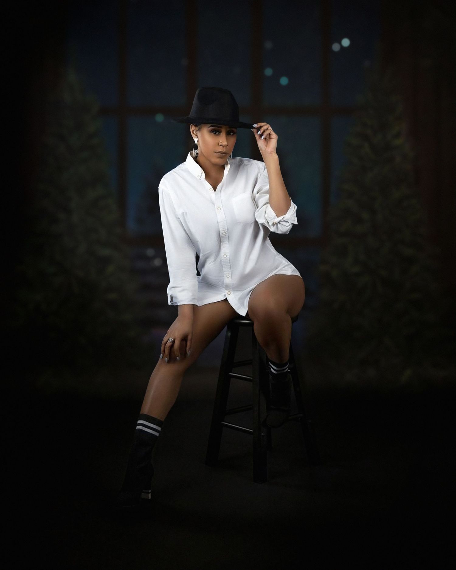 Artistic portrait of someone in a white button down shirt and black hat sitting on a stool against a dark background.