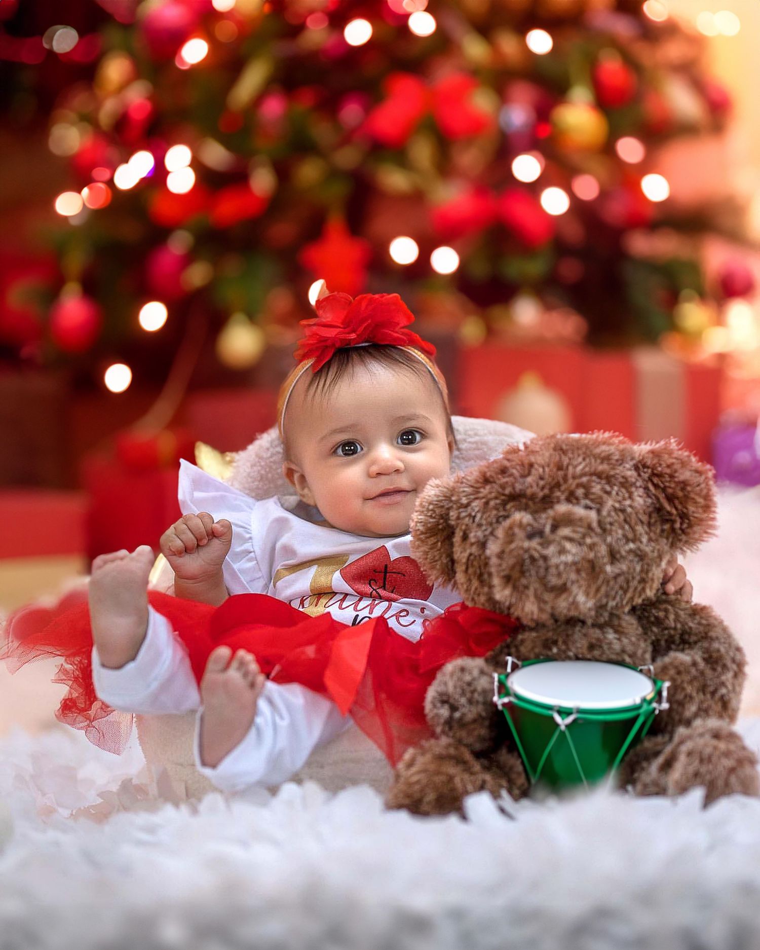 Baby in white outfit lying on red blanket with teddy bear and Christmas lights in background.