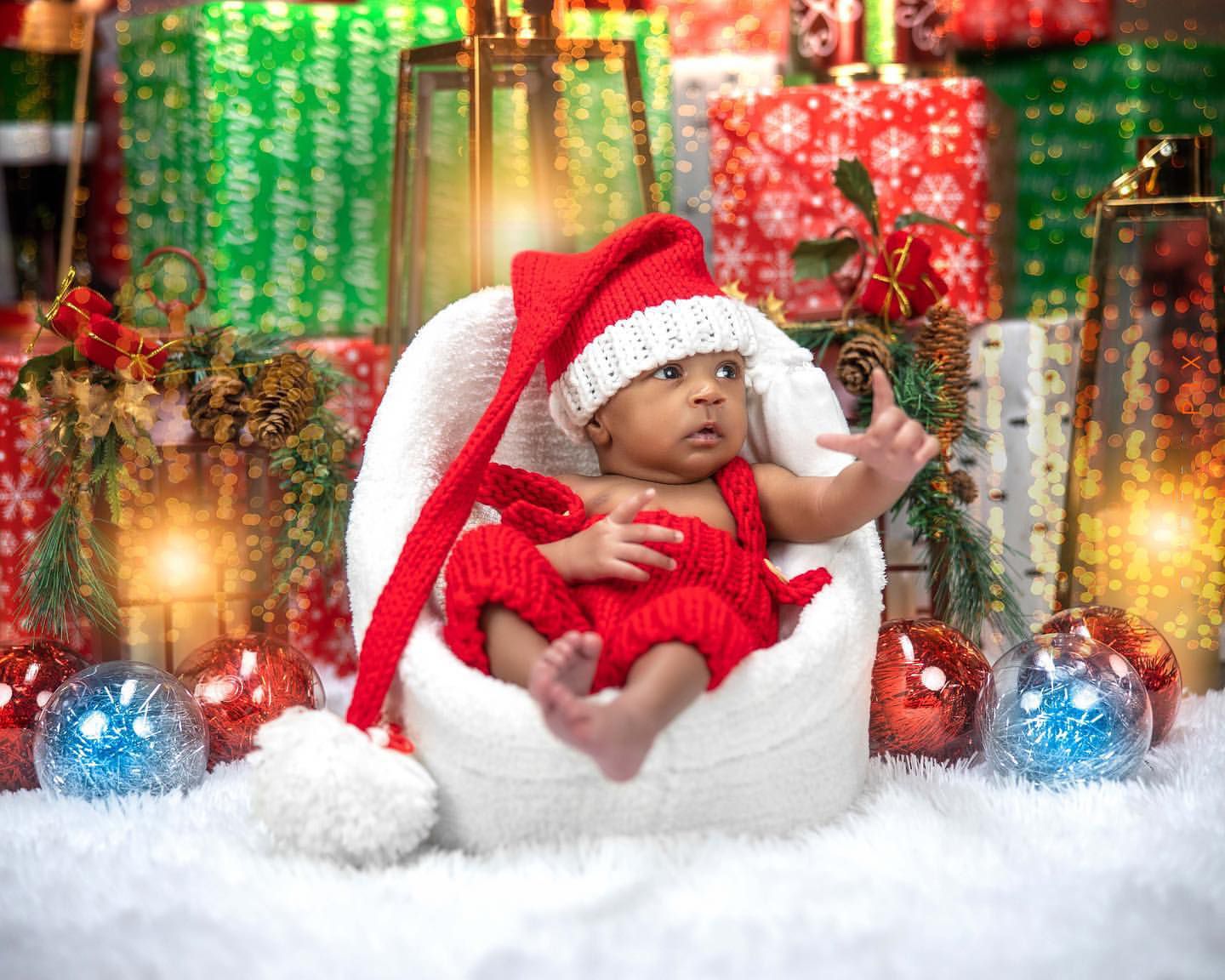Newborn baby wearing red Santa outfit posed in white eggshell chair surrounded by Christmas ornaments.