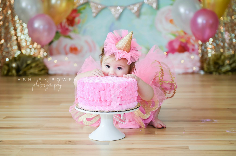 Baby girl in pink unicorn costume reaches for pink ruffled birthday cake at first birthday cake smash.