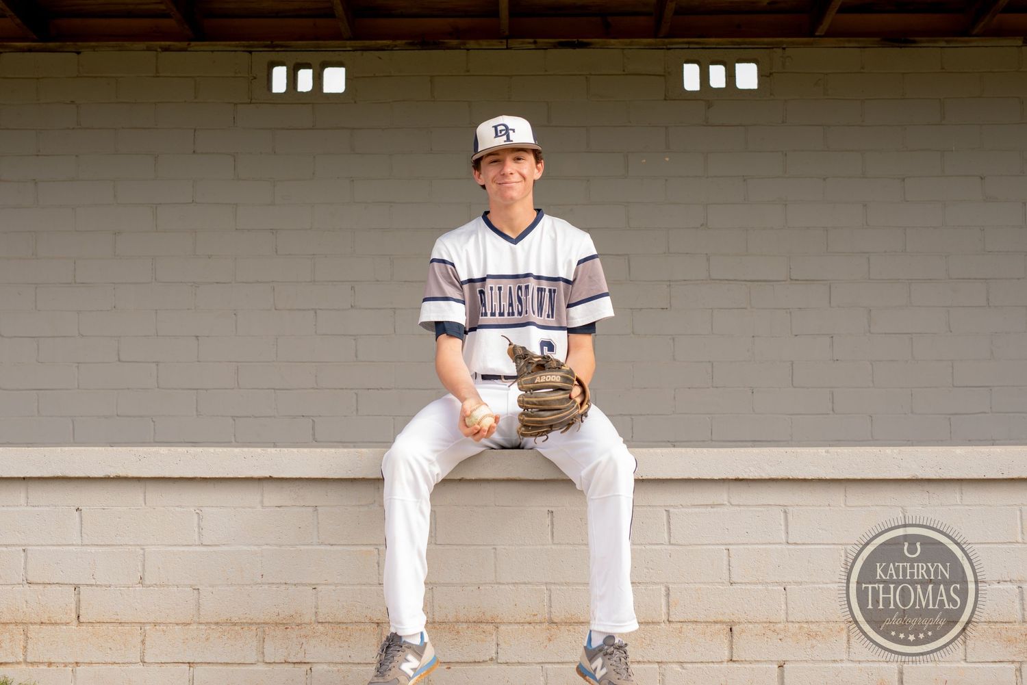 Baseball player in white uniform sits in concrete dugout with baseball glove.