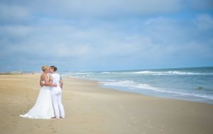 Couple in white attire embraces on empty beach with ocean waves in background.