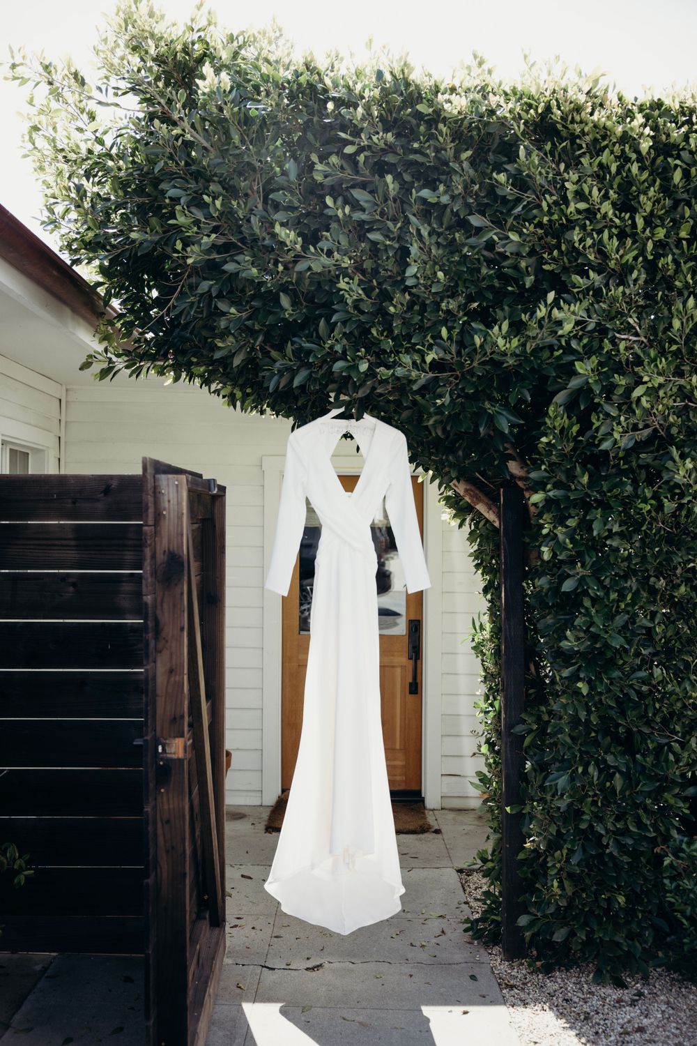 White wedding dress hanging outdoors against green foliage and white building exterior.