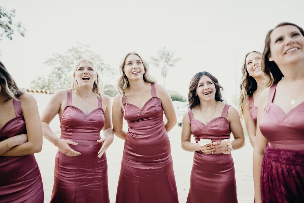 Bridesmaids wearing matching burgundy dresses pose together at an outdoor venue.