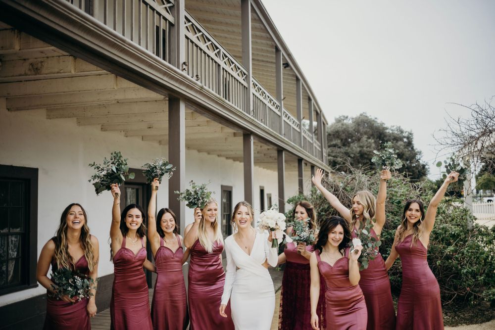 Wedding party celebrates with raised arms outside rustic building.
