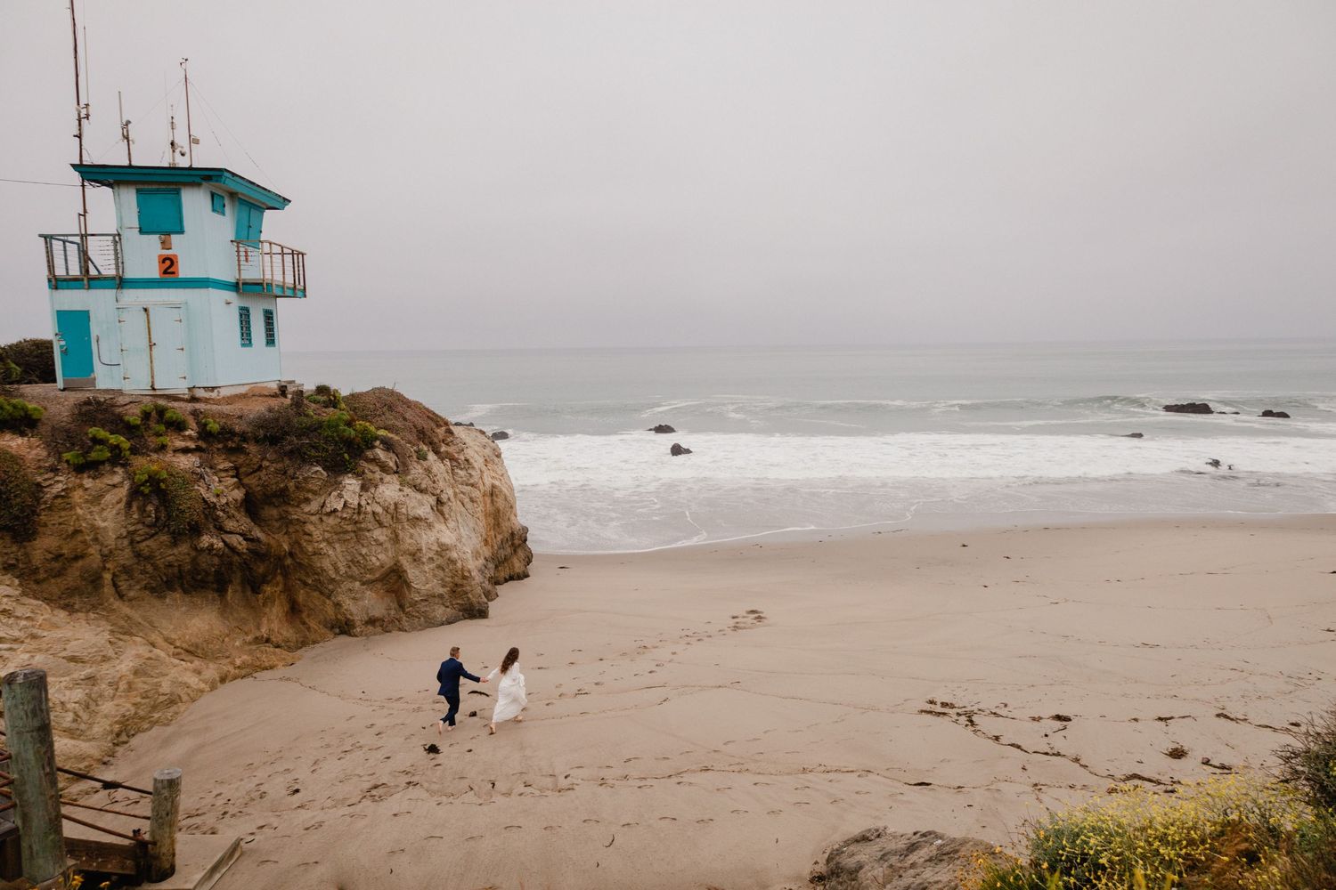 Sioux City, Iowa photographer. Bride and groom walking hand in hand on a misty beach near a blue lifeguard tower.
