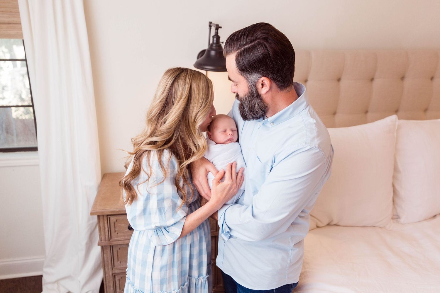 Parents in soft blue and white outfits share tender moment with newborn in bedroom setting.