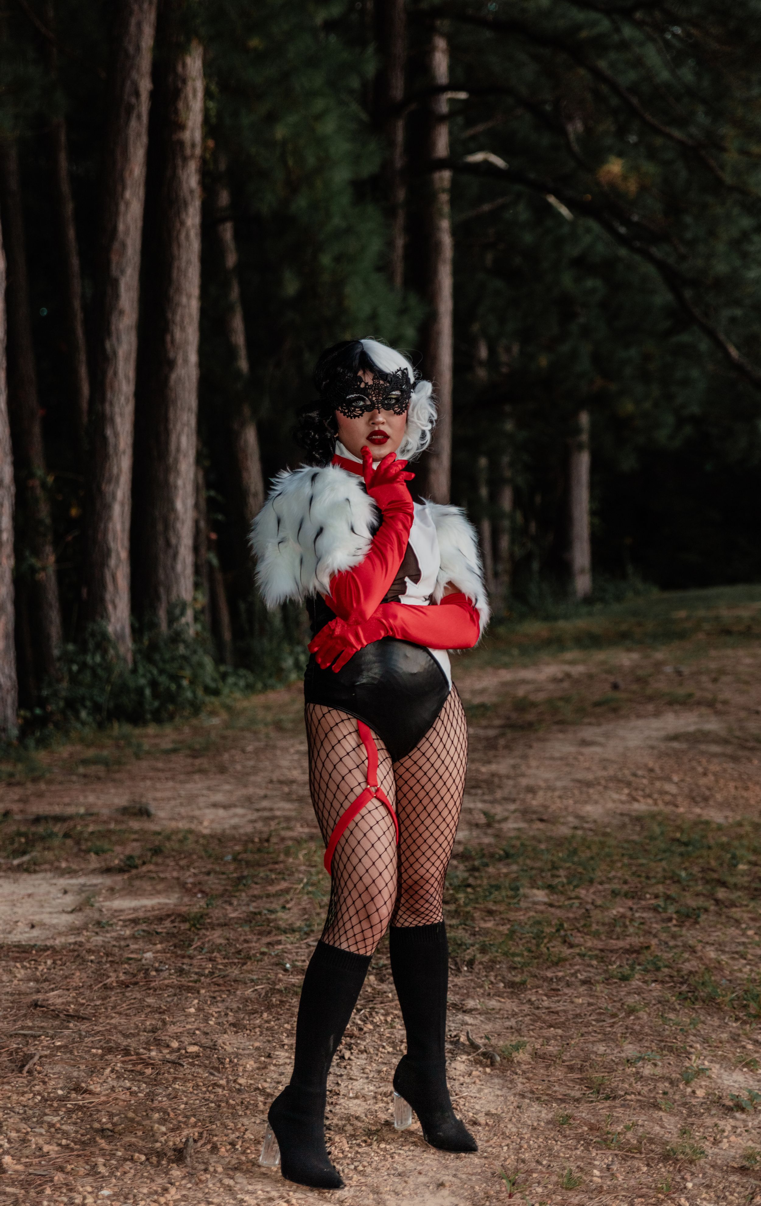 Cosplay photoshoot featuring a Cruella de Vil inspired costume with red gloves in a forest setting.