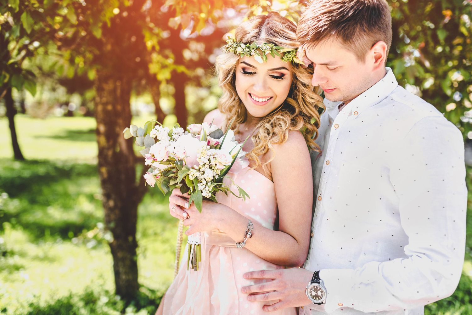 Romantic spring wedding photo with couple embracing in sunlit garden setting with floral bouquet.