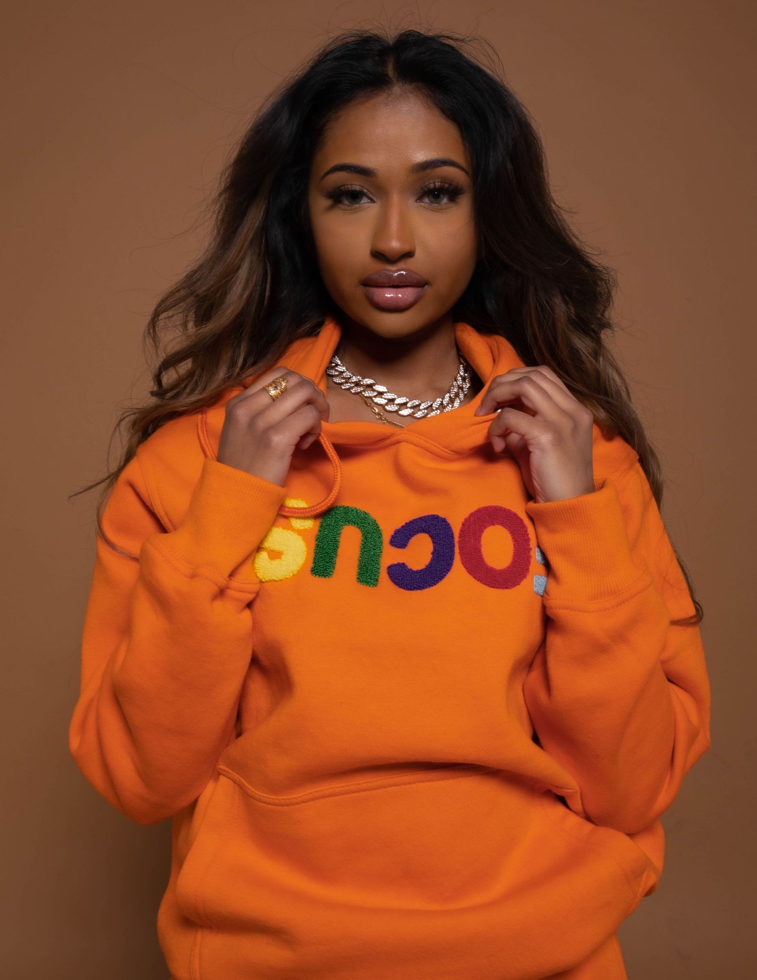 Orange sweatshirt with colorful text and silver chain necklace in studio portrait against brown background.