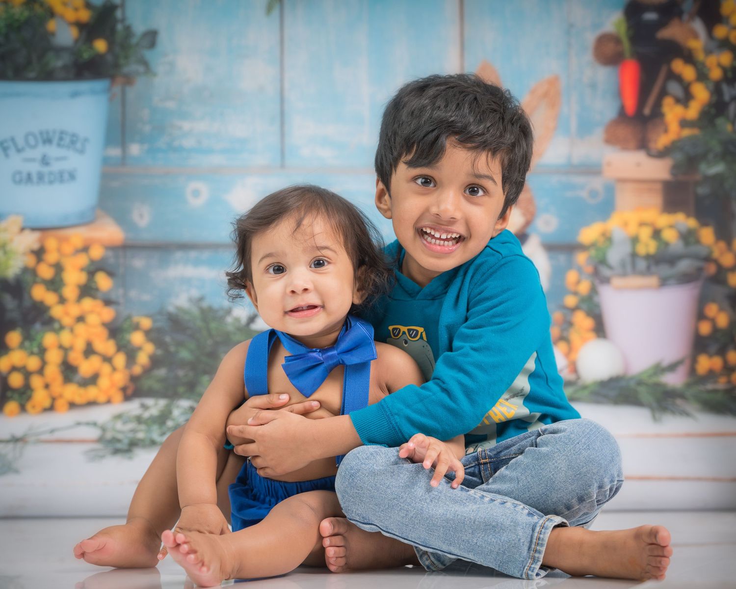Two young siblings in blue outfits pose together for a portrait against a rustic backdrop with yellow flowers.