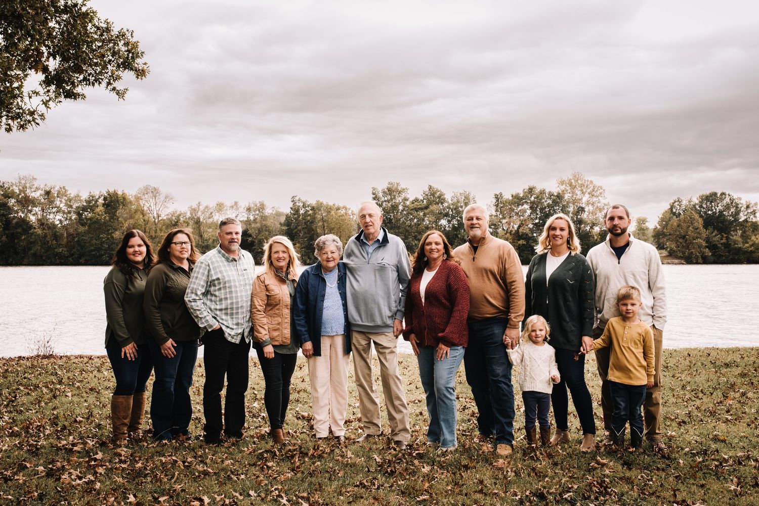 A large family group poses together outdoors in casual attire against a scenic autumn backdrop.
