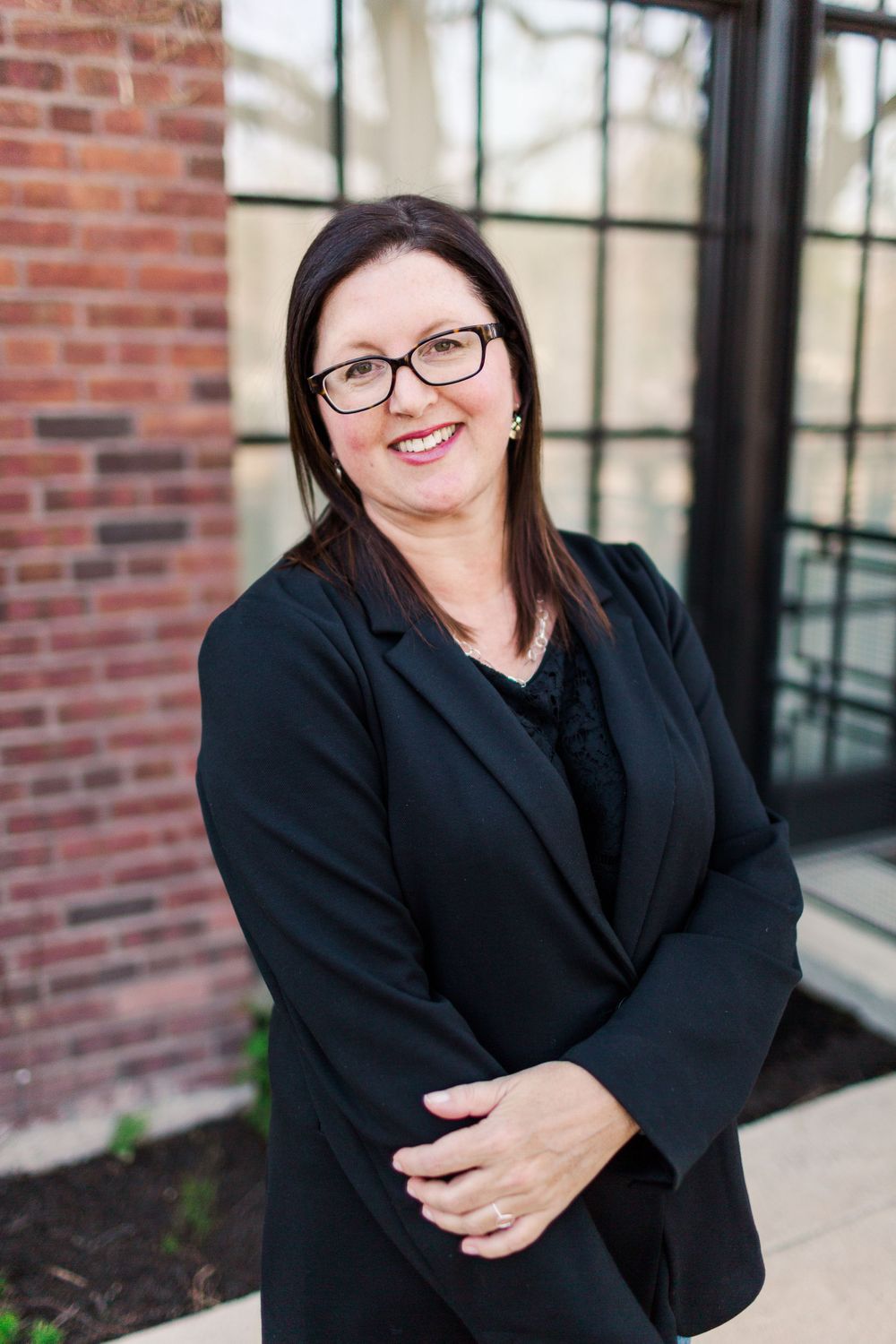 Business portrait of person in black blazer and glasses against brick wall background.