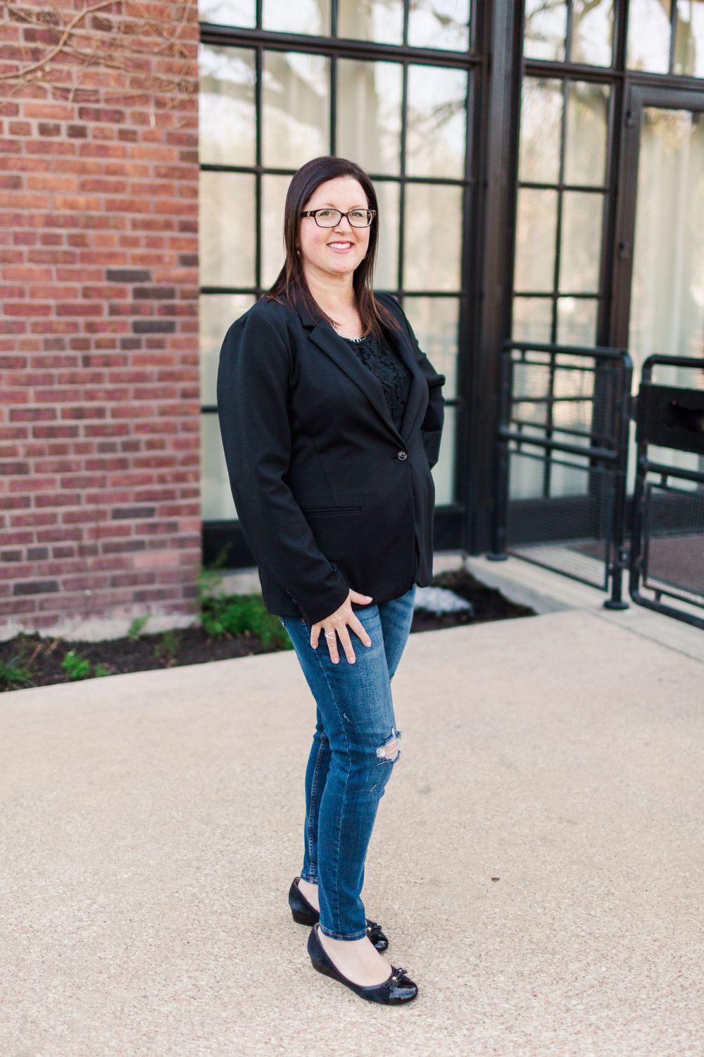 Full-length portrait of person in black blazer and ripped jeans against brick building.