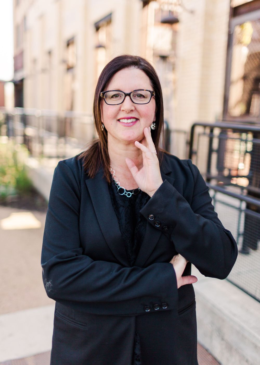 Professional headshot of person in black blazer and glasses against urban building backdrop.