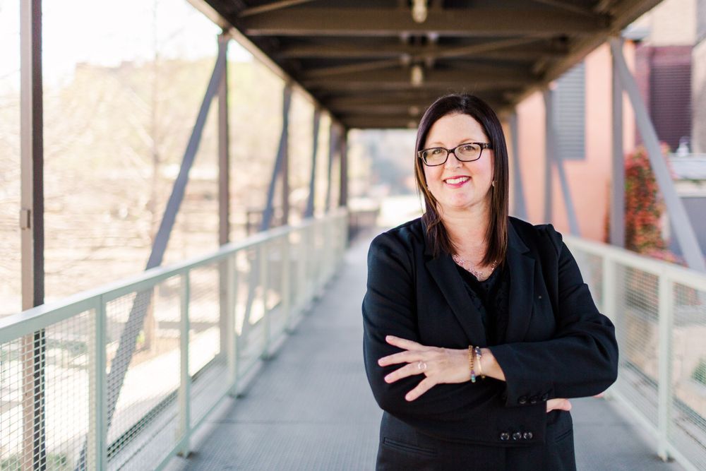 Professional portrait of person in black blazer on covered walkway with industrial architecture.