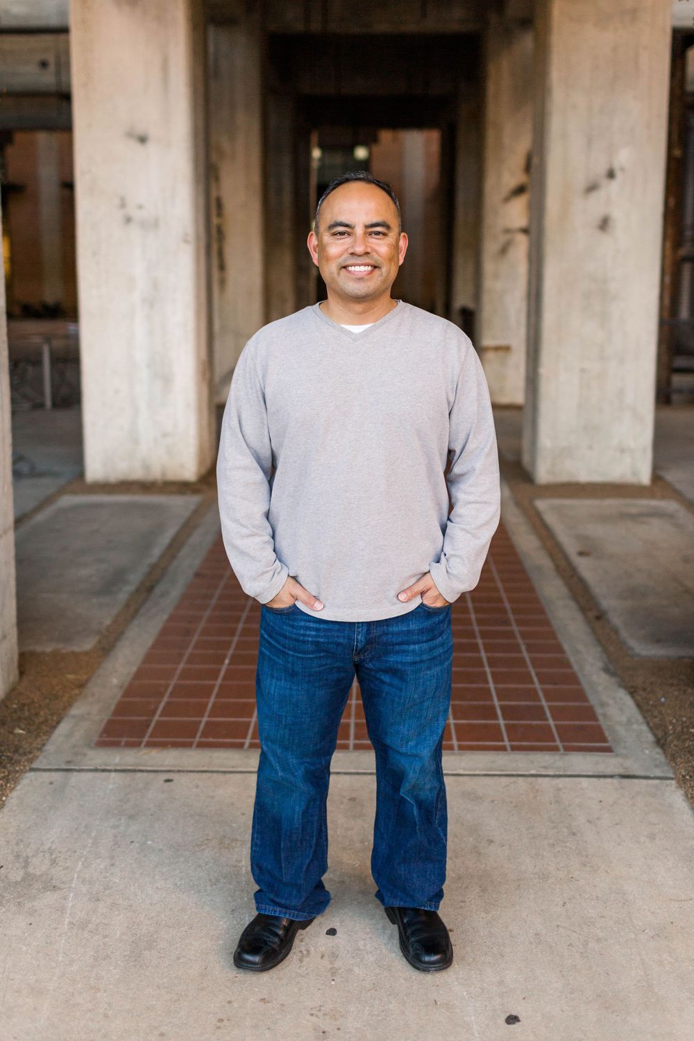 Professional portrait of person in grey sweater and jeans standing in front of a columned building entrance.