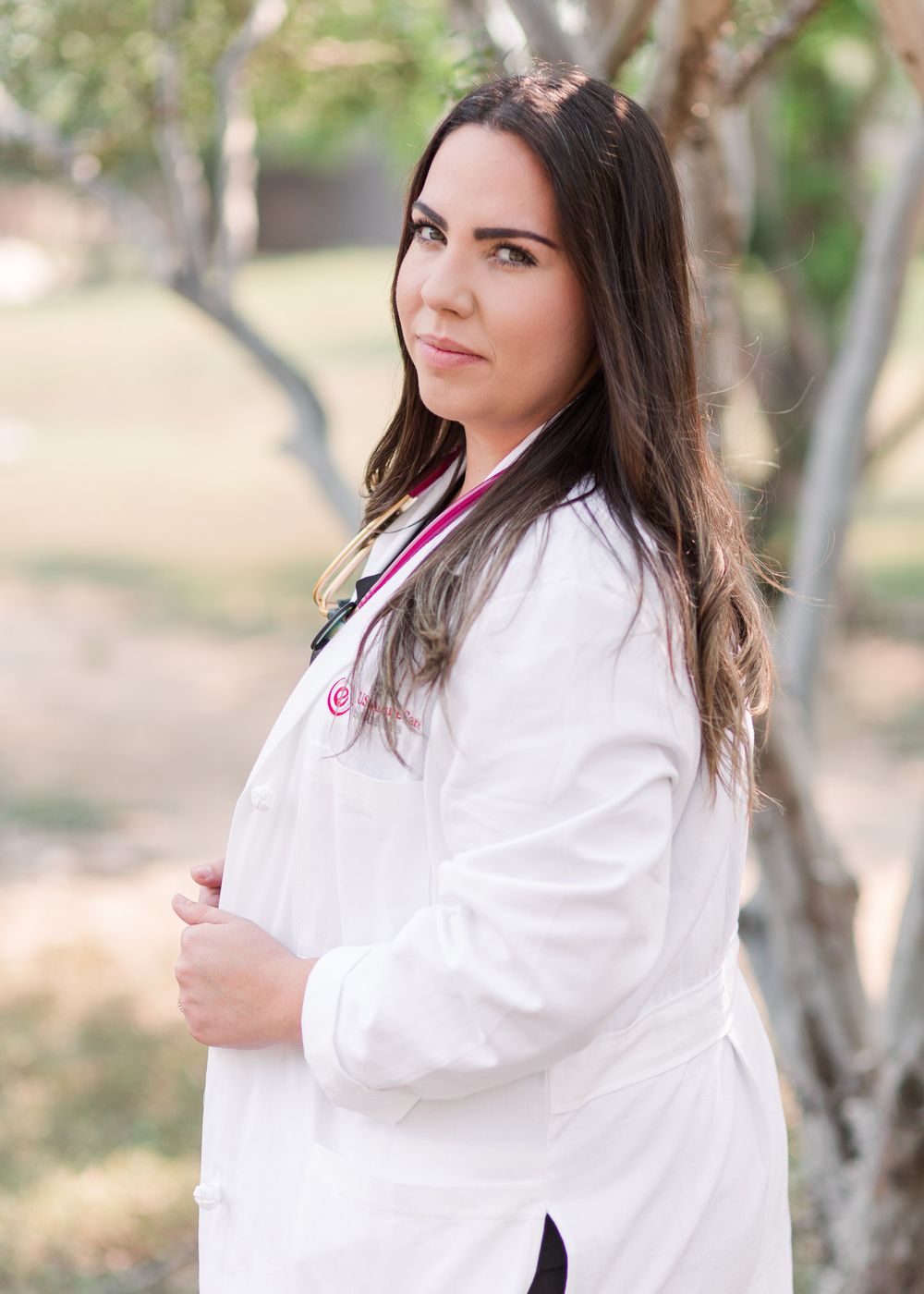 Healthcare professional in white coat posed against blurred natural background with trees.