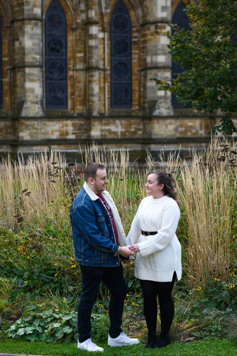 Couple standing among tall grass with gothic church windows in background.