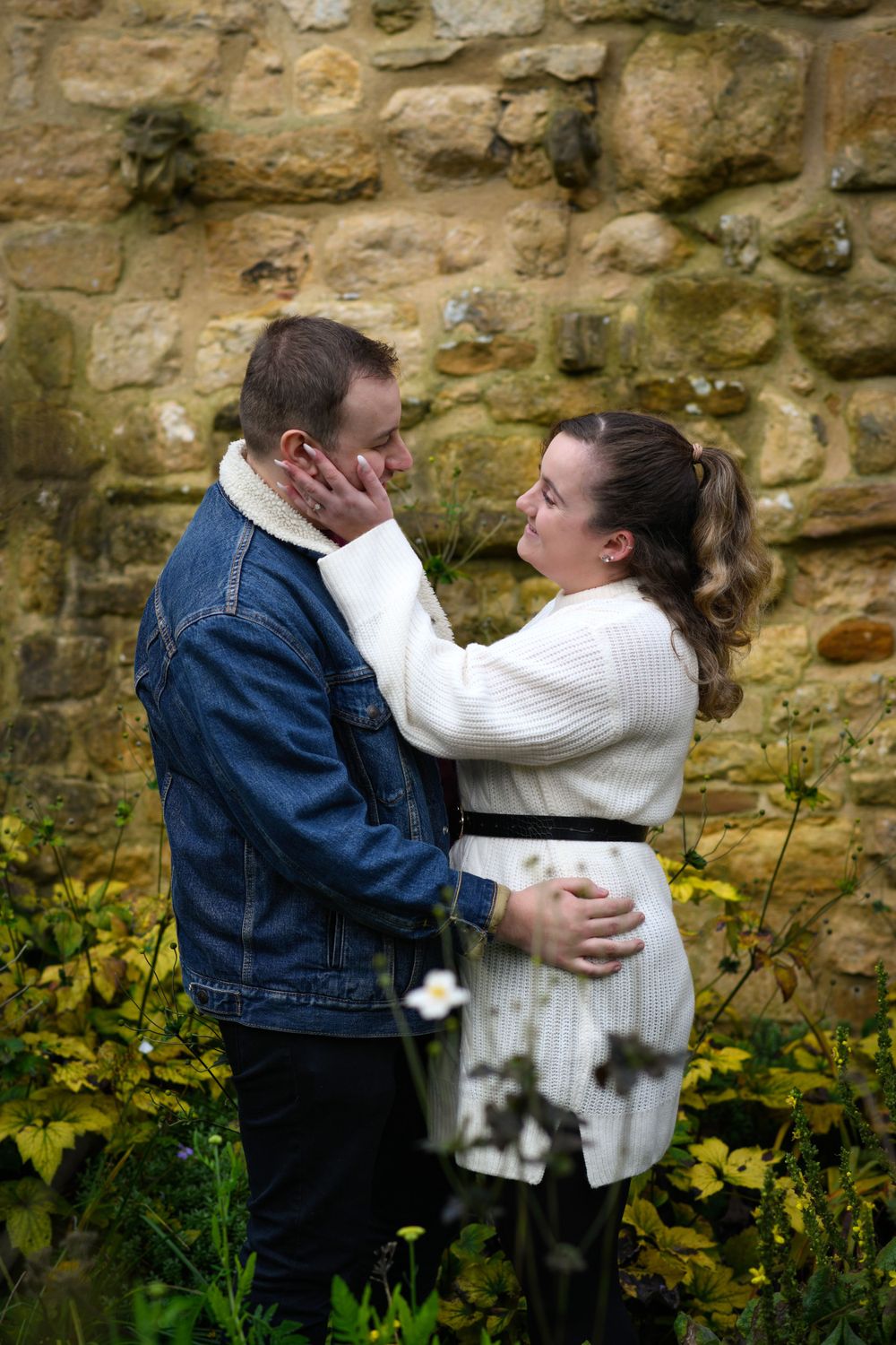 Intimate moment between couple in casual attire against stone wall with yellow flowers below.