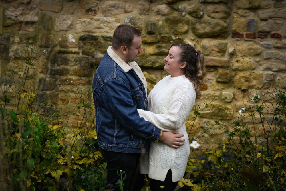 A romantic couple embraces against a rustic stone wall background with wildflowers.