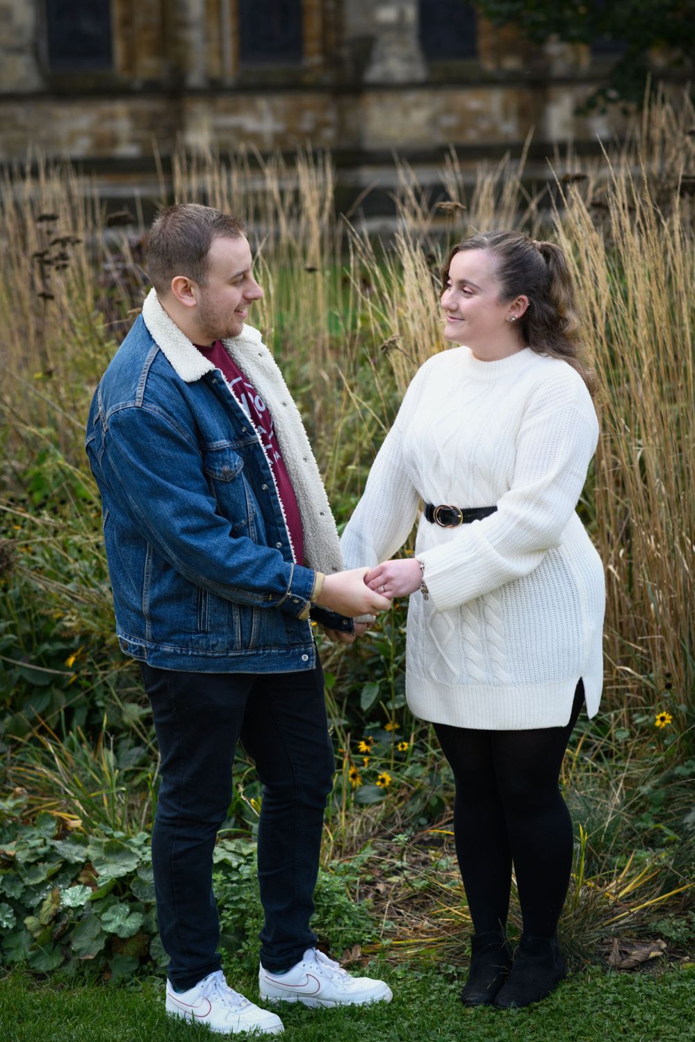 Couple holds hands in front of stone church with tall grass in foreground.