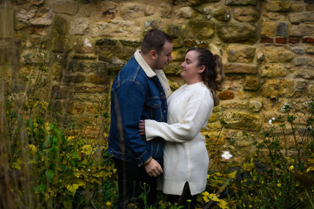 Romantic couple embraces against weathered stone wall surrounded by wildflowers.