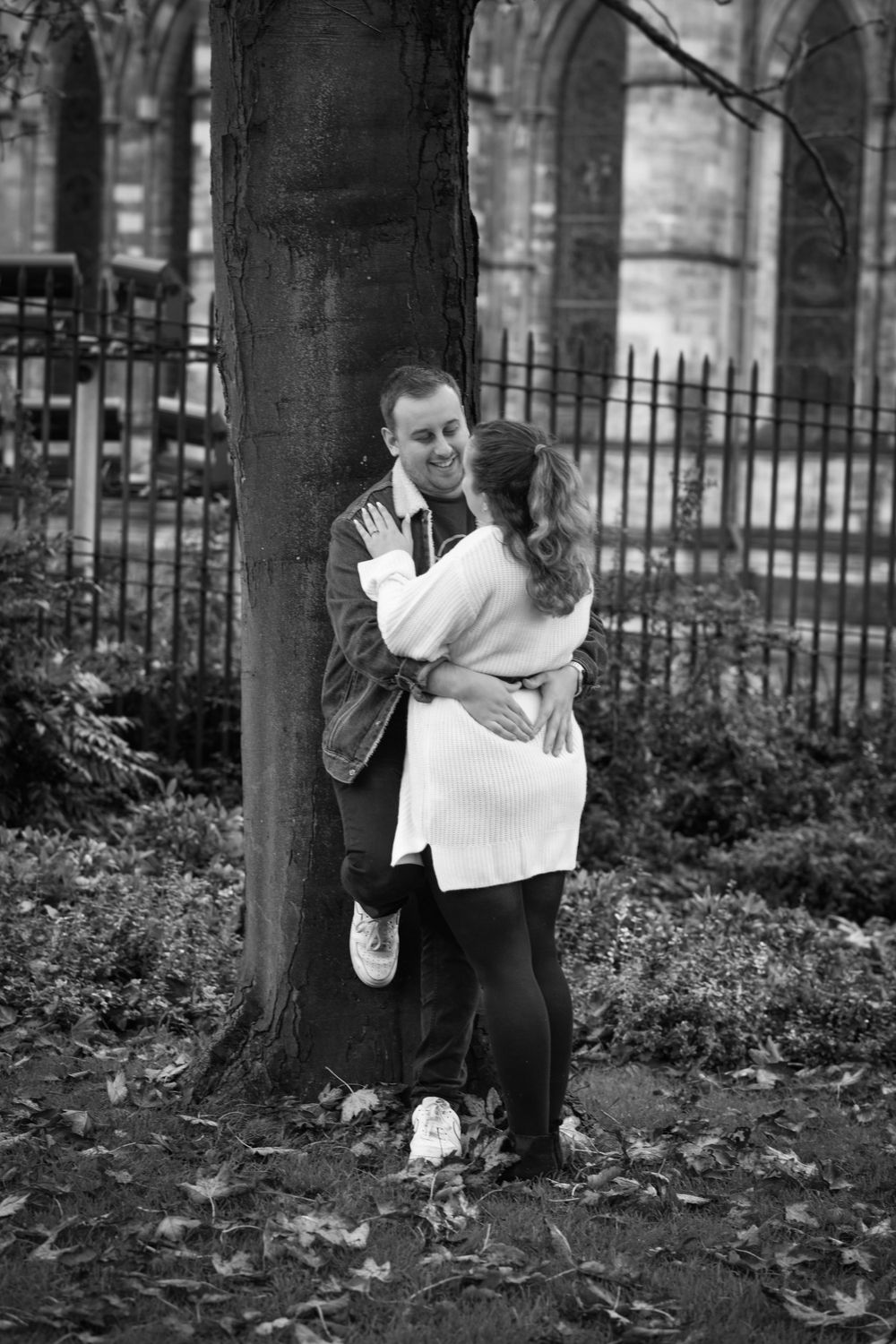 Black and white portrait of couple embracing by tree trunk with gothic architecture in background.