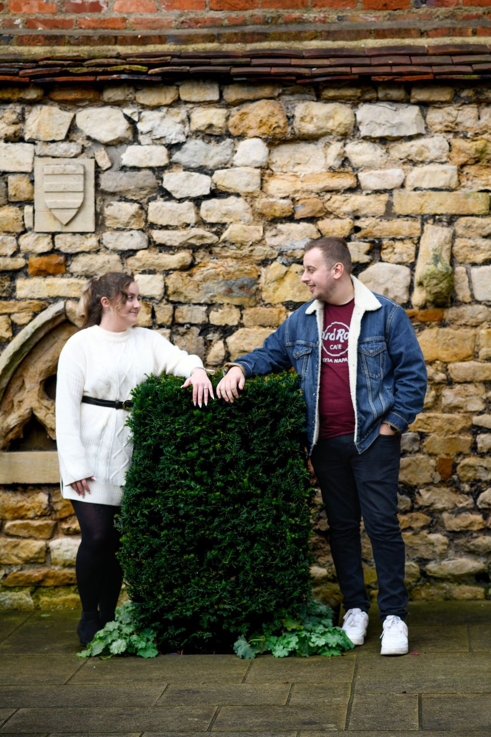 Couple poses playfully with a large hedge bush against historic stone wall architecture.