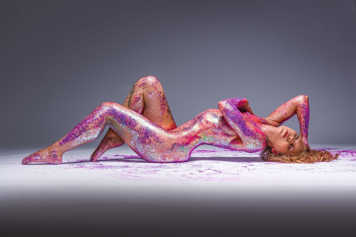 Artistic nude covered in pink and purple glitter powder on grey background.