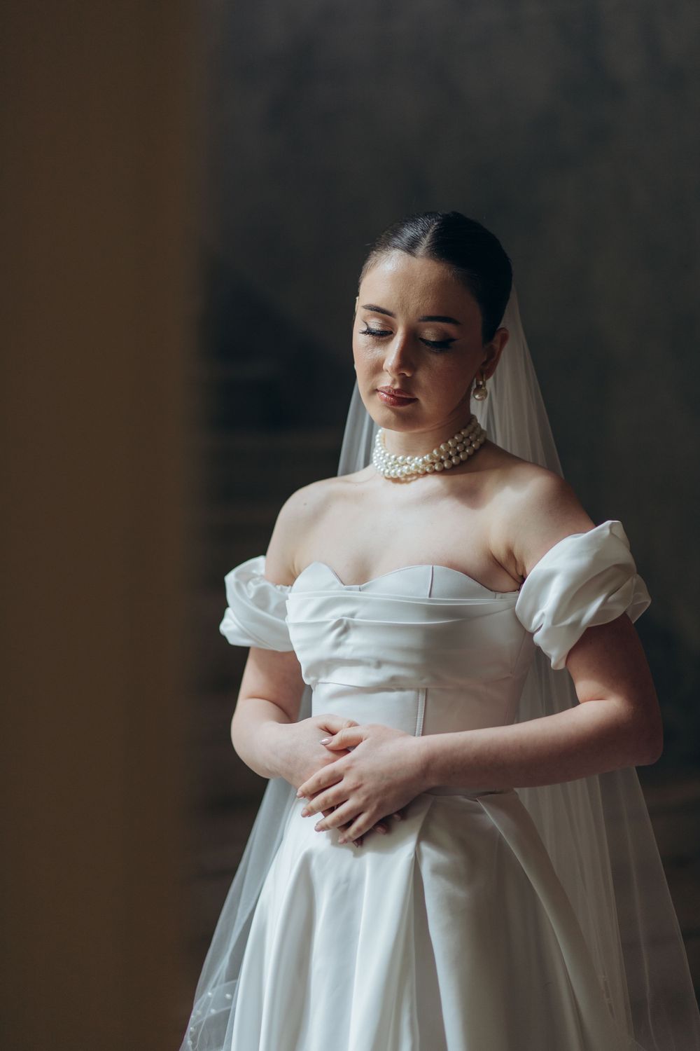 Elegant bride in off-shoulder gown with puff sleeves and pearl choker in window light.