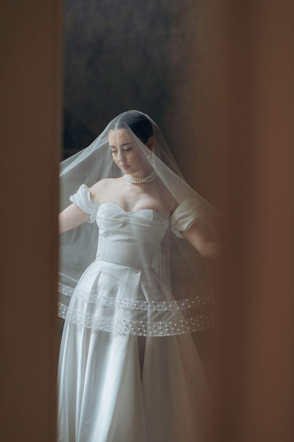 Dreamy bridal portrait through doorframe showing detailed lace veil and structured gown.
