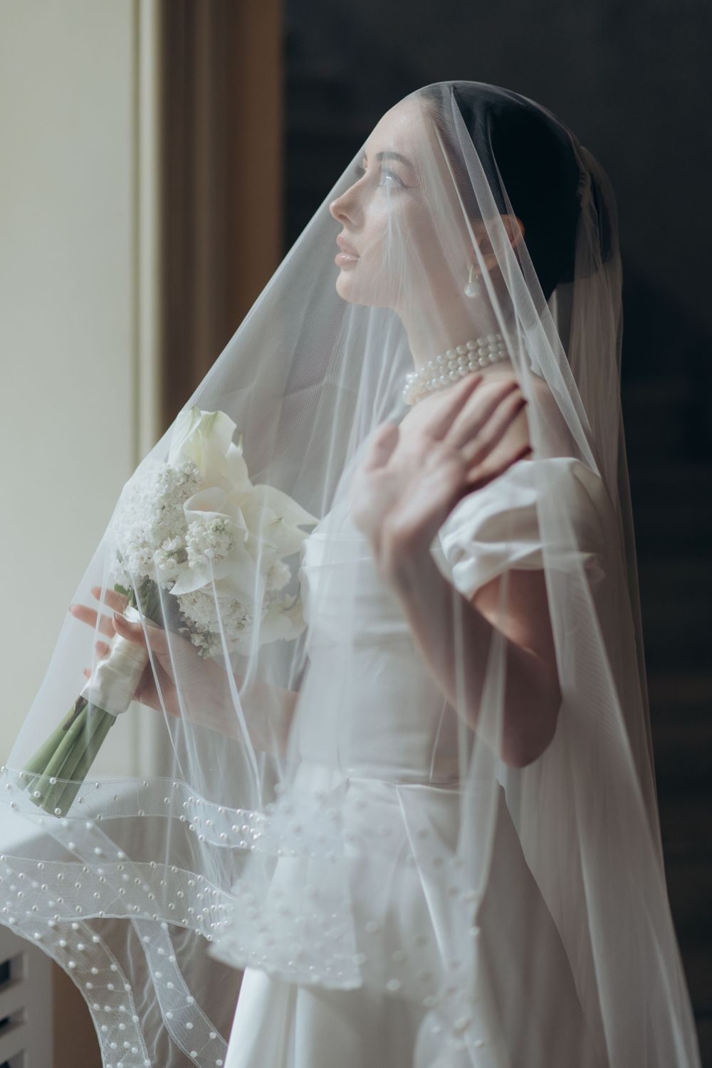 Ethereal bridal portrait with lace veil and white bouquet in soft window light.