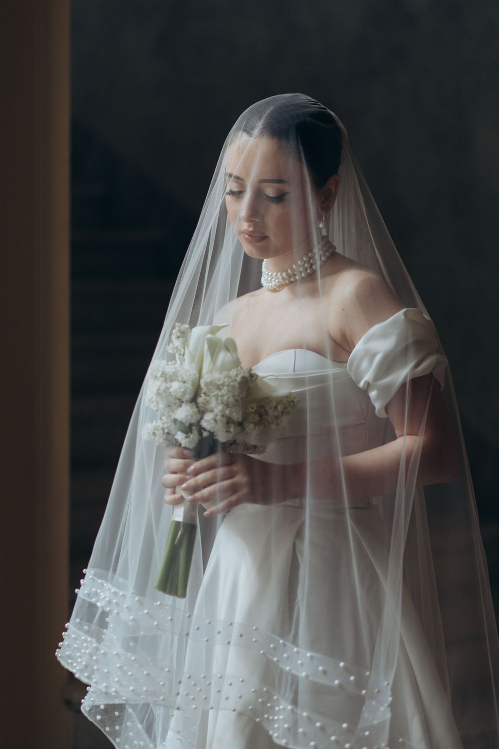 Elegant bride in white strapless gown and veil holds bouquet of white flowers against dark background.