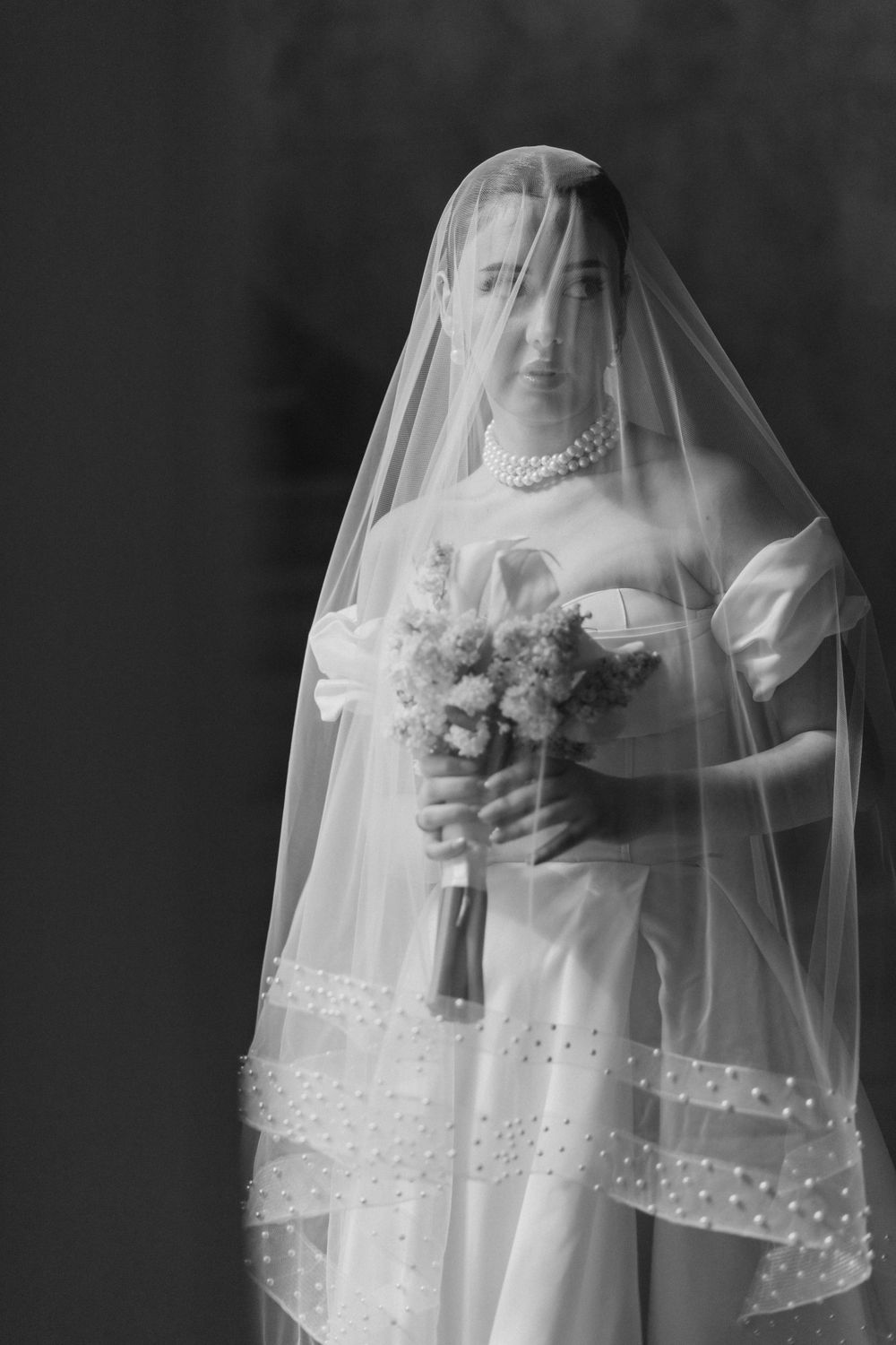 Black and white portrait of bride wearing pearl necklace and ornate veil holding floral bouquet.