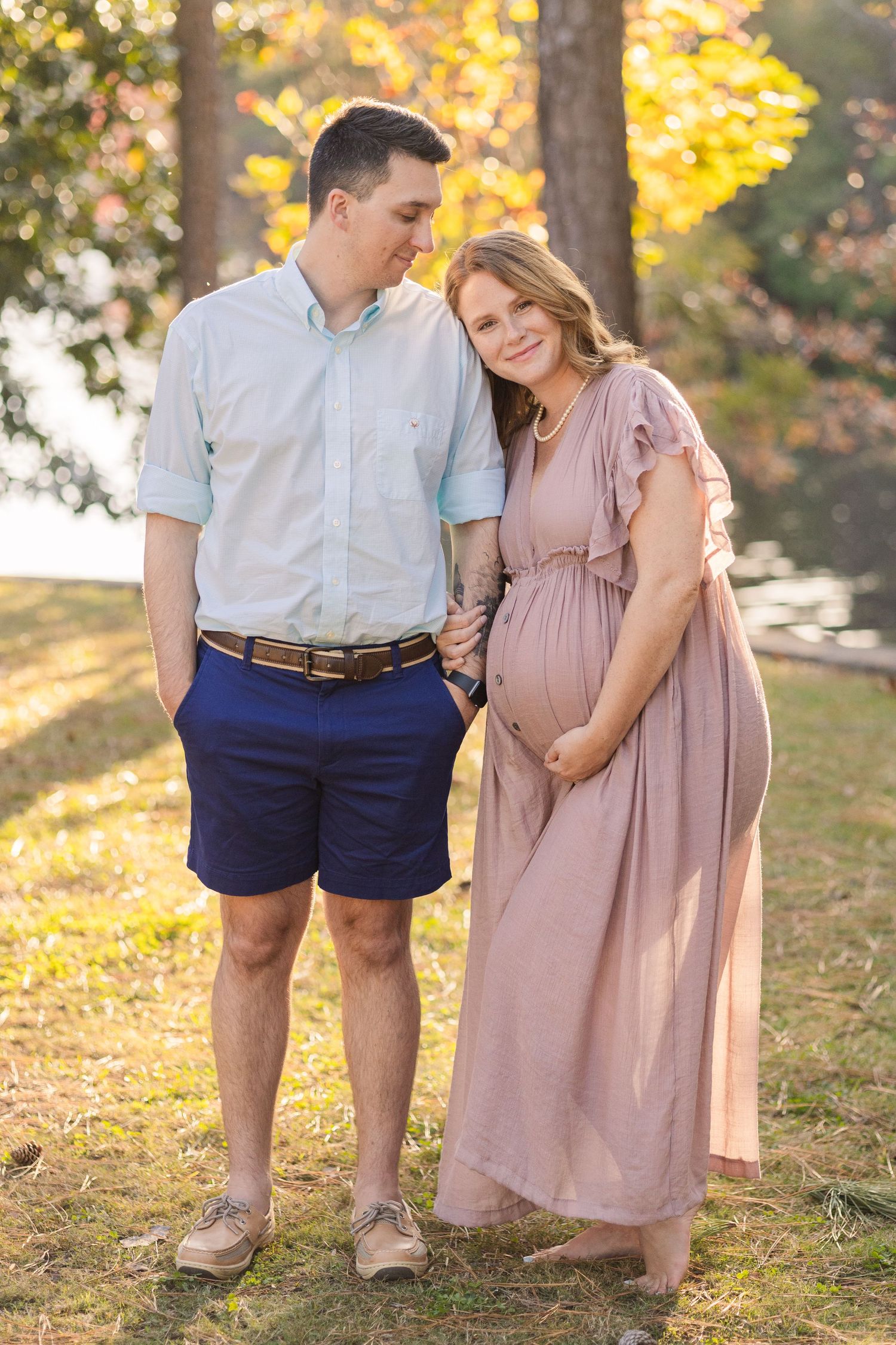 A tender maternity portrait captured in a park with fall colors and warm sunlight.
