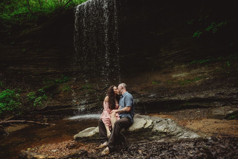 A couple shares an intimate moment while sitting on rocks near a gentle waterfall.