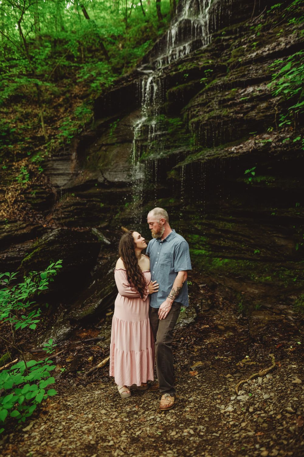 A couple shares an intimate moment near a small waterfall in a wooded area.